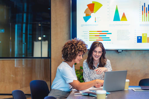 Two women looking at a laptop with charts on the wall