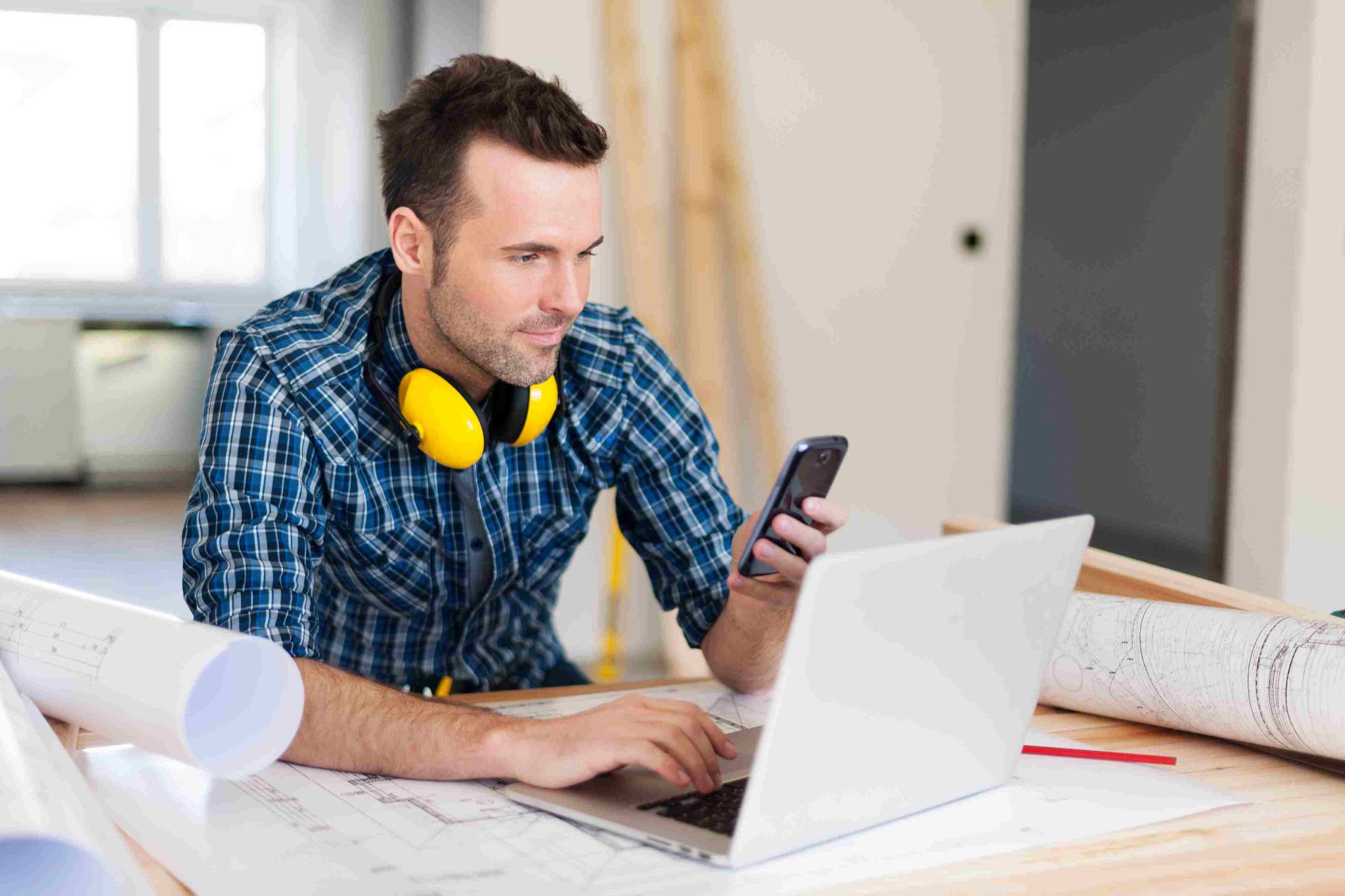 Construction worker with yellow earmuffs looking at phone near laptop