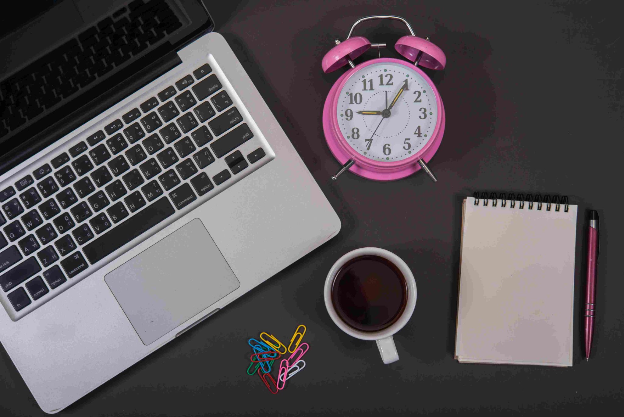 Laptop, pink alarm clock, coffee cup, and notebook on a desk