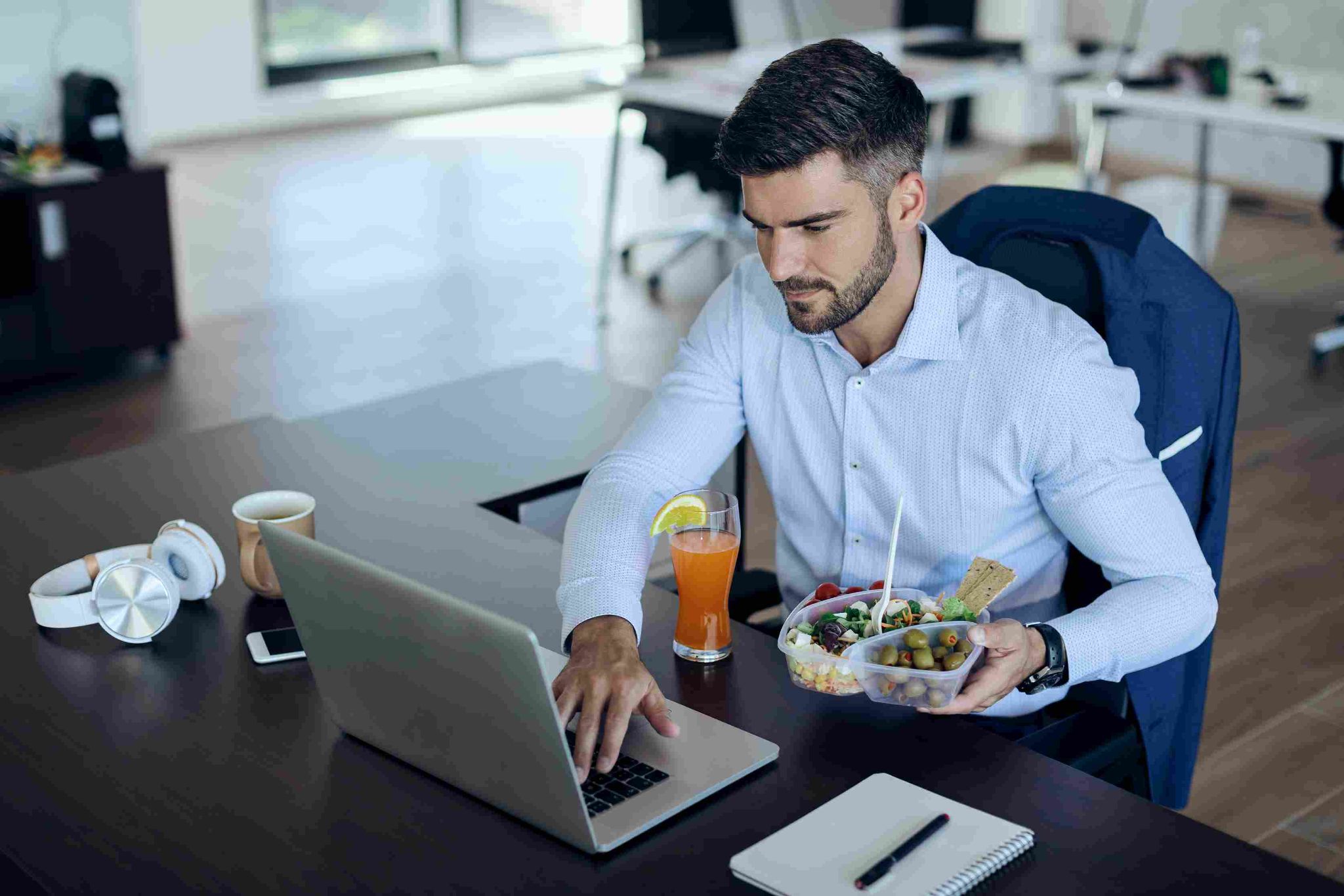 Man sitting at a desk eating a salad while looking at a laptop