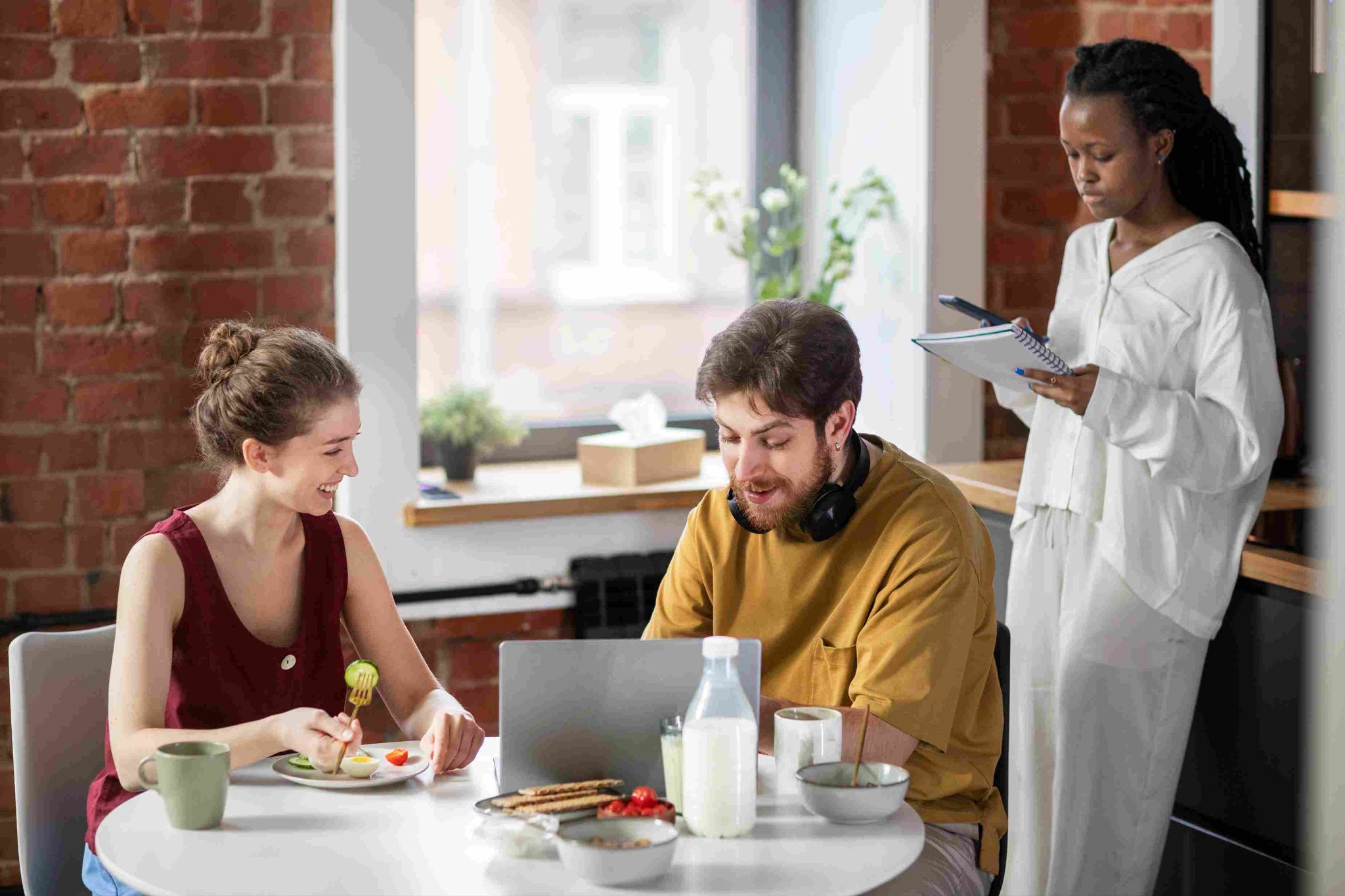 Three people in an office setting, two sitting at a table with food and laptops, one standing holding a notepad