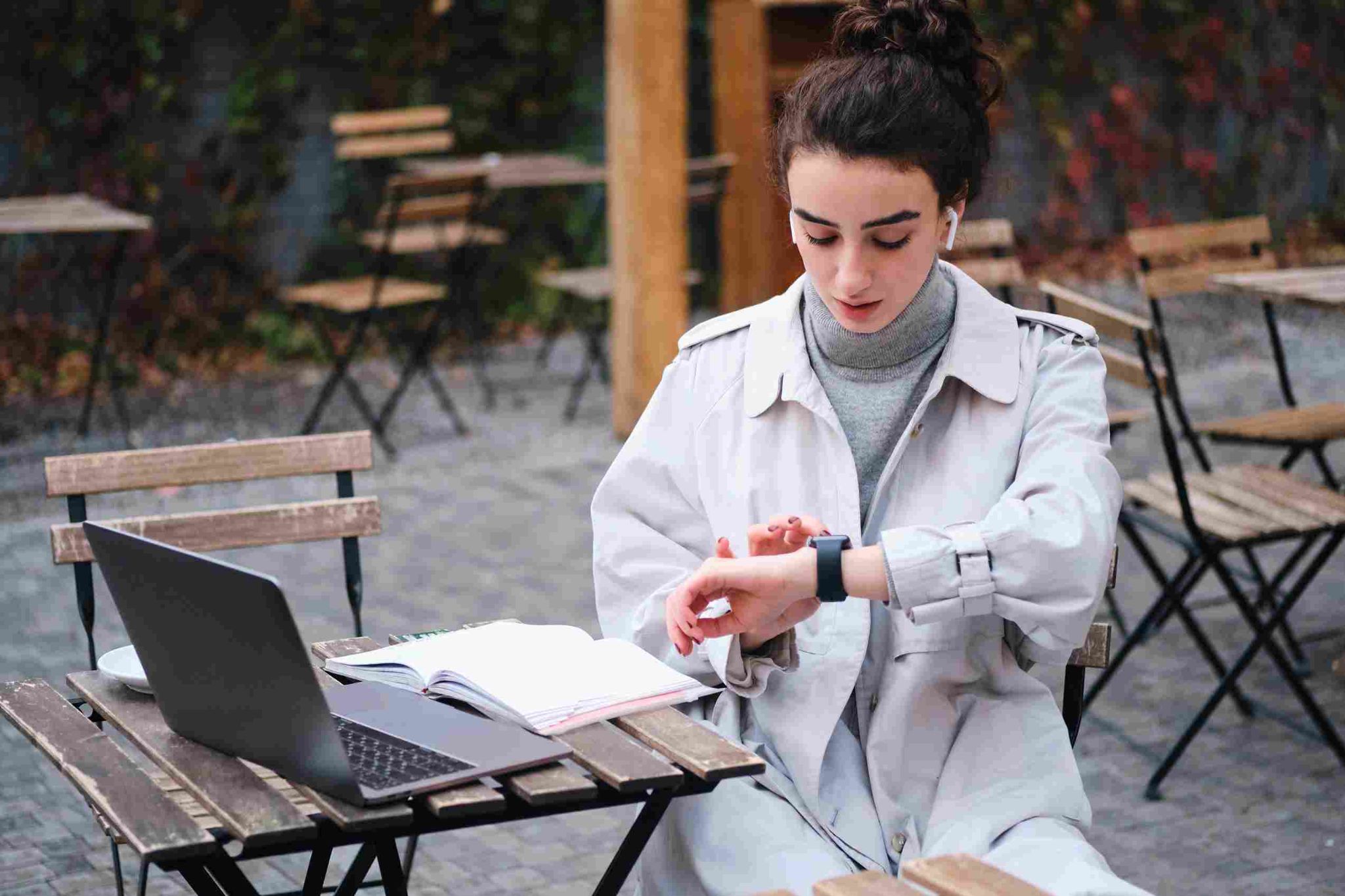 Woman looking at a smartwatch while working at an outdoor cafe table with a laptop
