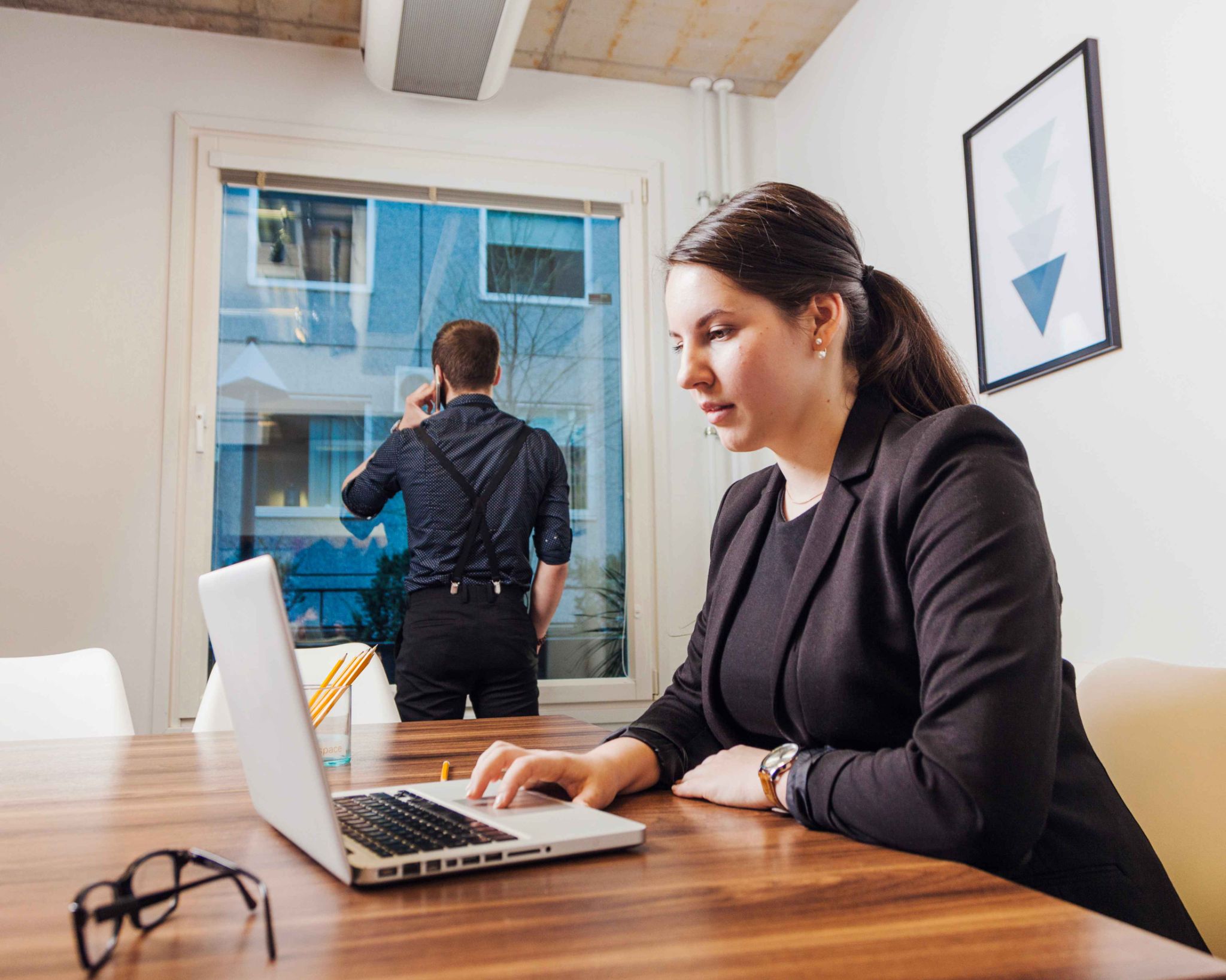 Woman working on a laptop with a man on the phone in the background