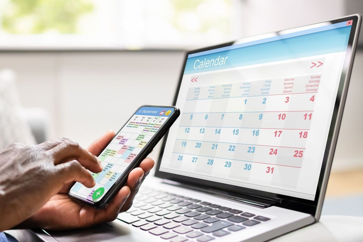 Person holding a smartphone with a calendar interface on a laptop behind it