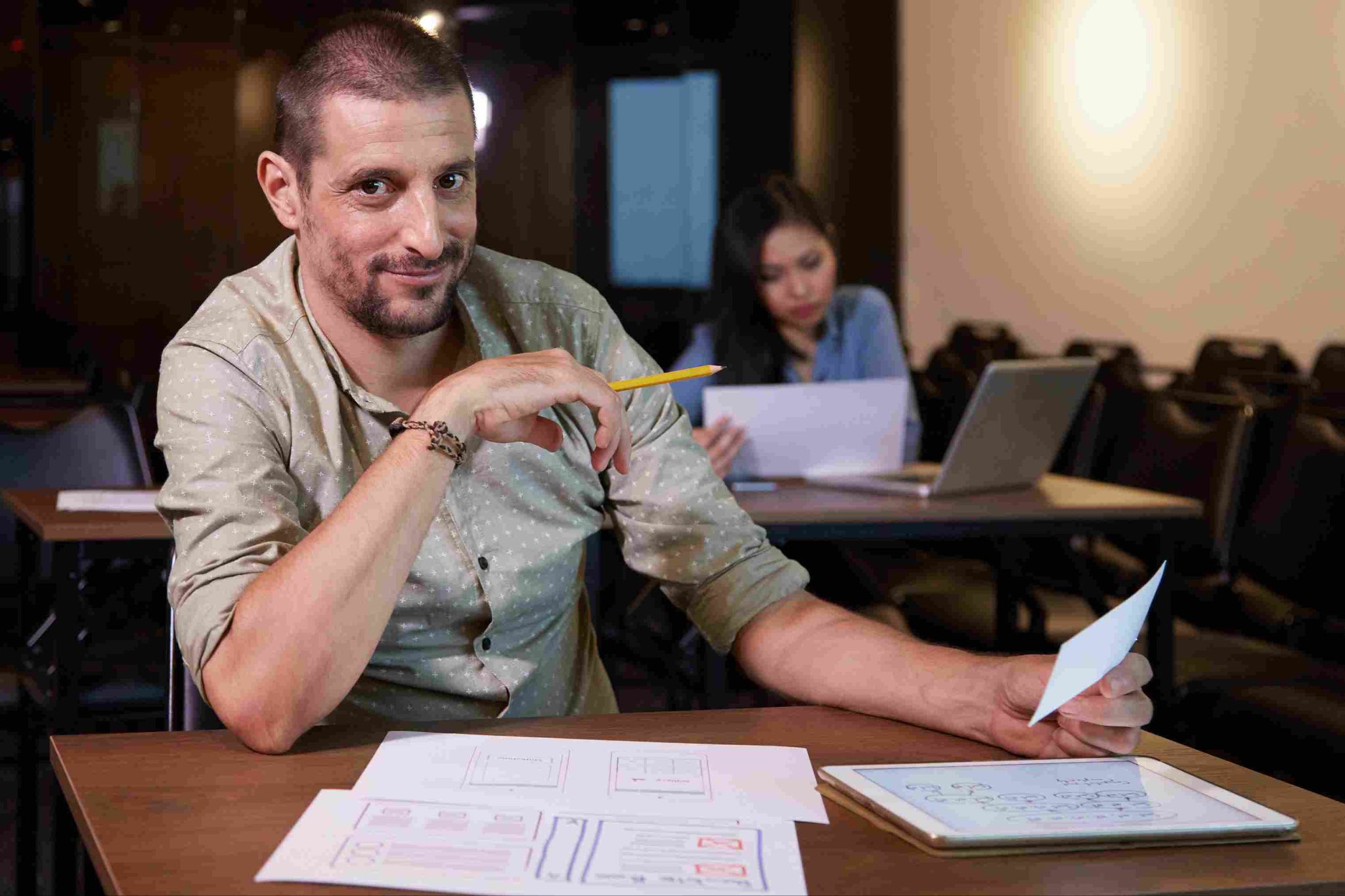 Man sitting at a desk reviewing documents with a woman working in the background