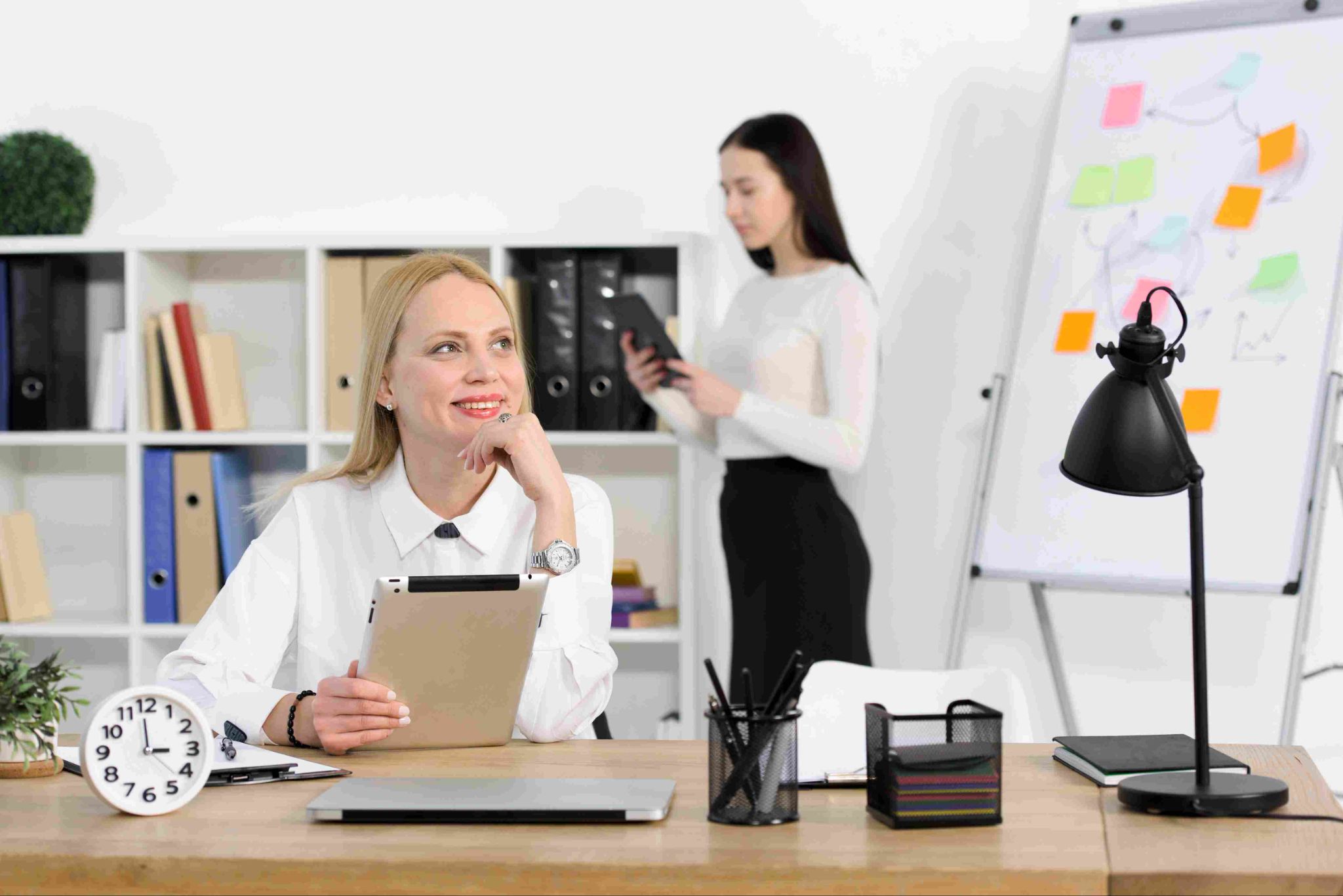 Woman sitting at a desk with another woman in the background by a whiteboard
