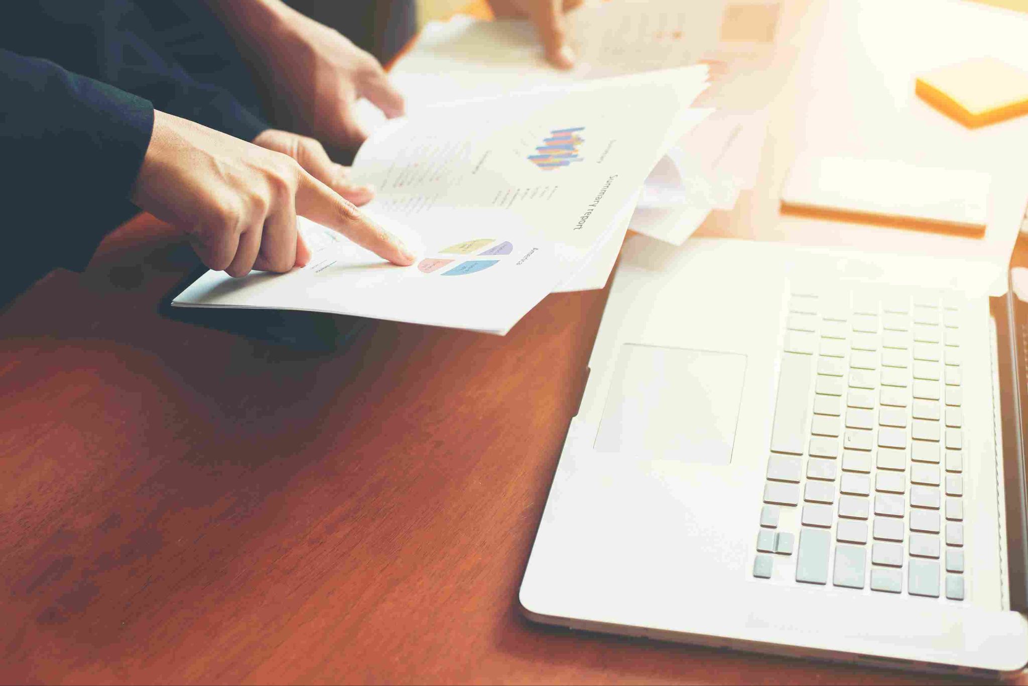 Hands pointing at charts/papers on a desk next to a laptop