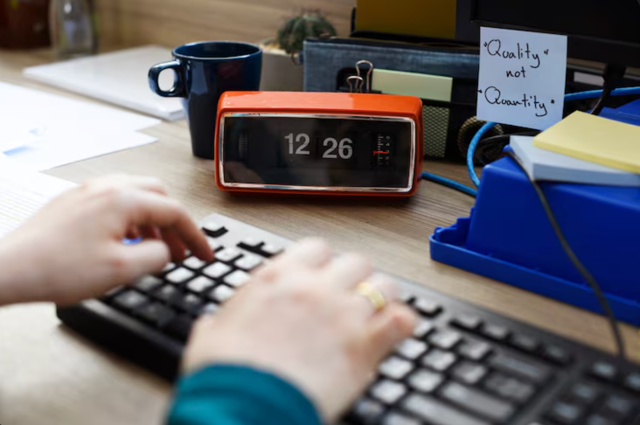 Person typing on laptop next to a digital clock
